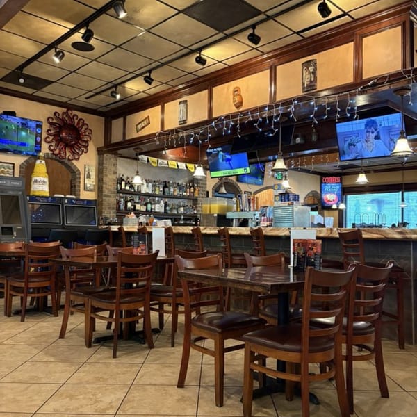 Interior of a restaurant with empty tables, chairs, a bar, and multiple TVs showing sports and other content.