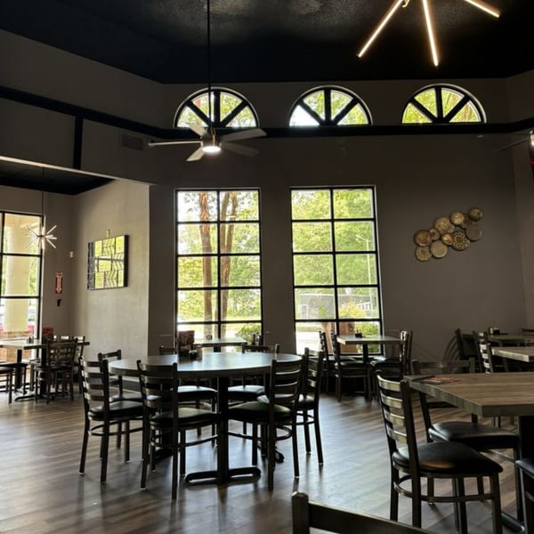 Spacious restaurant dining room with dark tables and chairs, large windows, and decorative lighting.
