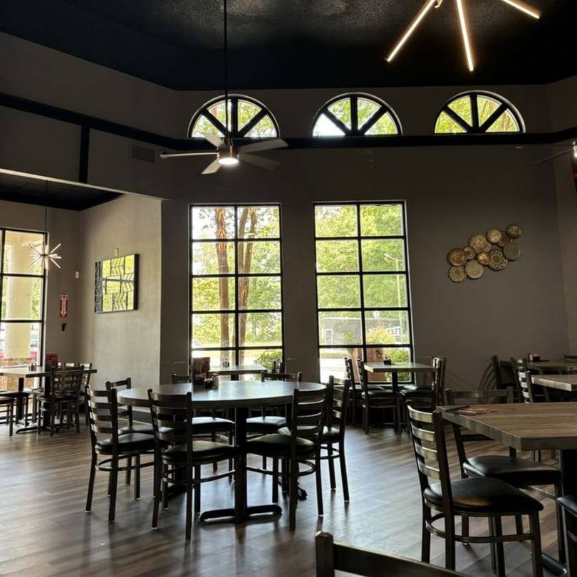 Spacious restaurant dining room with dark tables and chairs, large windows, and decorative lighting.