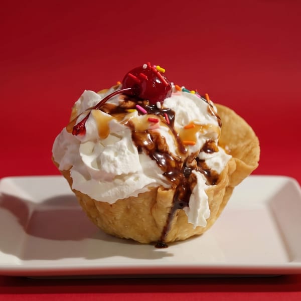 Dessert taco shell filled with whipped cream, chocolate, caramel, sprinkles, and a cherry on a white plate.