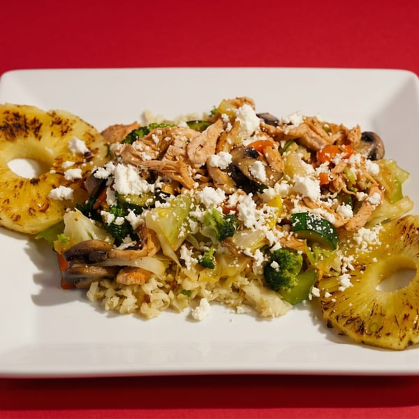 Chicken and vegetable stir-fry with crumbled feta cheese and grilled pineapple slices on a white plate.